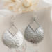 Close-up of sterling silver pear-shaped hammered dangle earrings with French hook ear wires on a marble surface.

